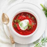 A bowl of red borscht soup topped with sour cream and fresh dill sits on a white plate next to a vintage spoon and sprigs of dill.