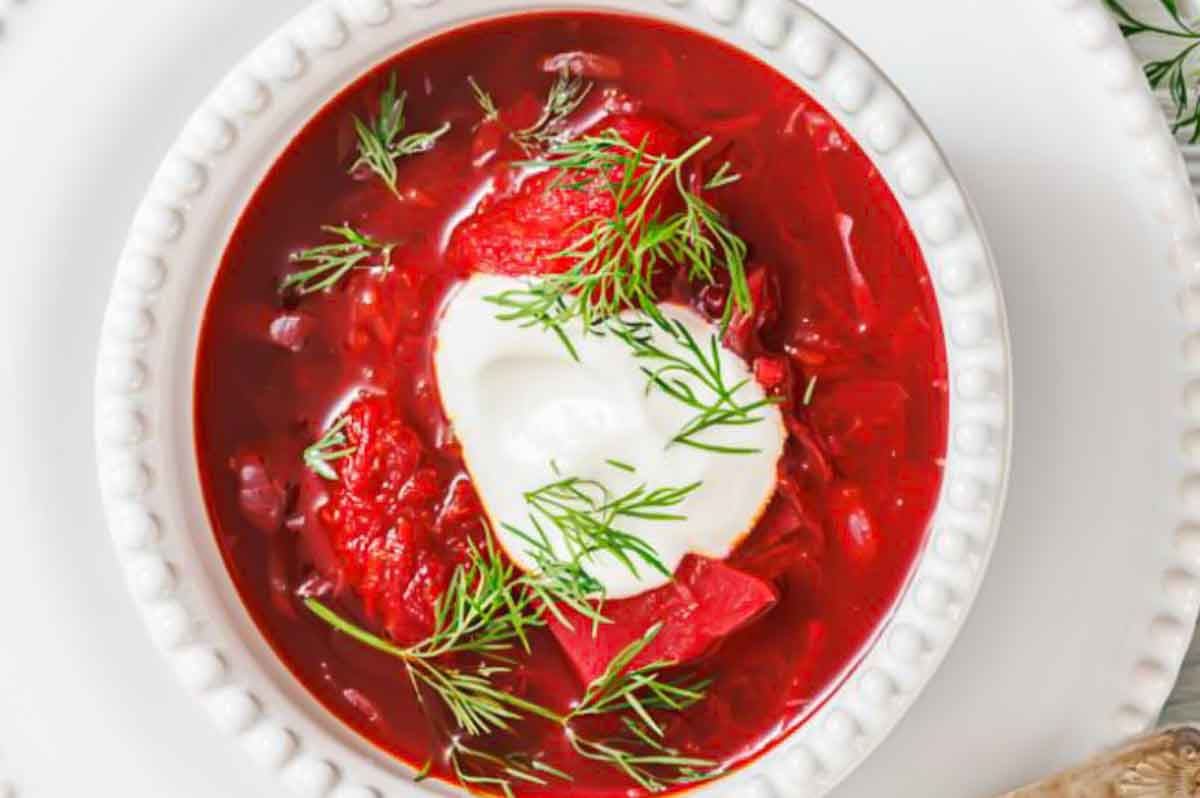 A bowl of red borscht soup topped with a dollop of sour cream and fresh dill, served in a white textured bowl.