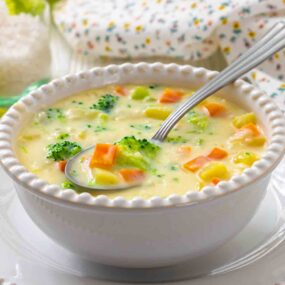A white bowl filled with creamy vegetable soup containing broccoli, carrots, and potatoes, with a spoon resting inside.
