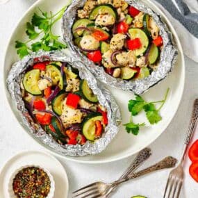 Two foil packets filled with baked chicken, zucchini, red bell peppers, and onions on a white plate, garnished with fresh parsley.