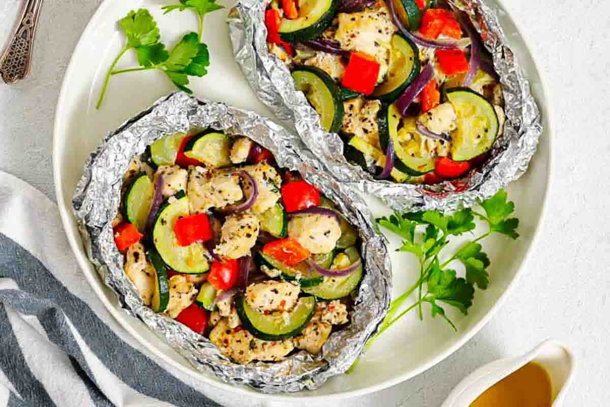 Two foil packets filled with baked chicken, zucchini, red bell pepper, and herbs on a white plate with parsley garnish.