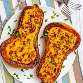 Two roasted butternut squash halves on a white plate, topped with chopped herbs and nuts, placed on a striped tablecloth with two forks beside the plate.