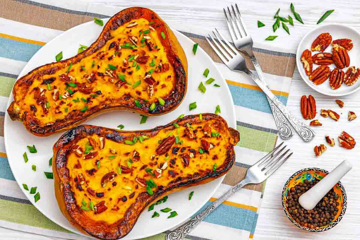 Two halves of roasted butternut squash with pecans and chopped herbs on a white plate, next to forks, pepper, and extra pecans on a striped tablecloth.