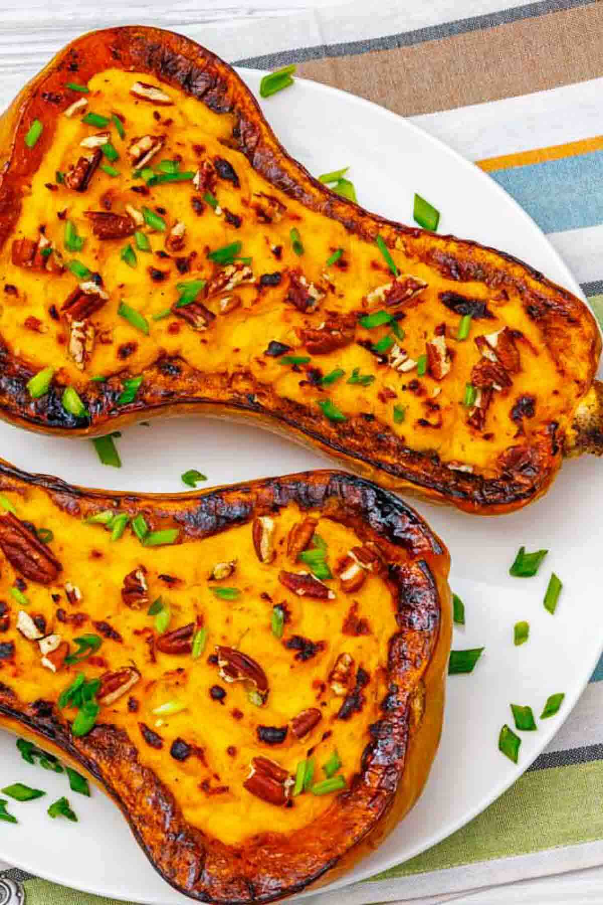 Two halves of roasted butternut squash topped with chopped pecans and green onions are served on a white plate.