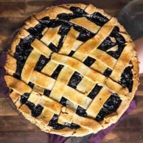 A freshly baked blueberry pie with a golden lattice crust is shown from above, resting on a wooden surface.