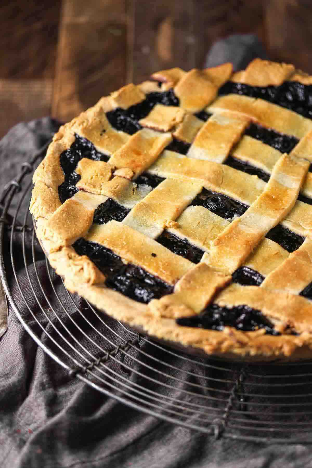 A lattice-topped blueberry pie sits on a cooling rack over a dark cloth and wooden surface.