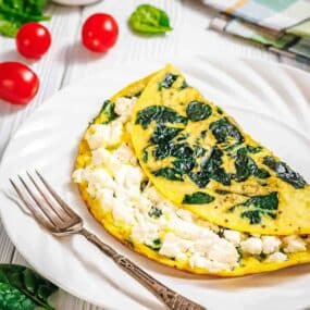 A spinach and feta cheese omelette is served on a white plate with a fork, surrounded by cherry tomatoes and spinach leaves.