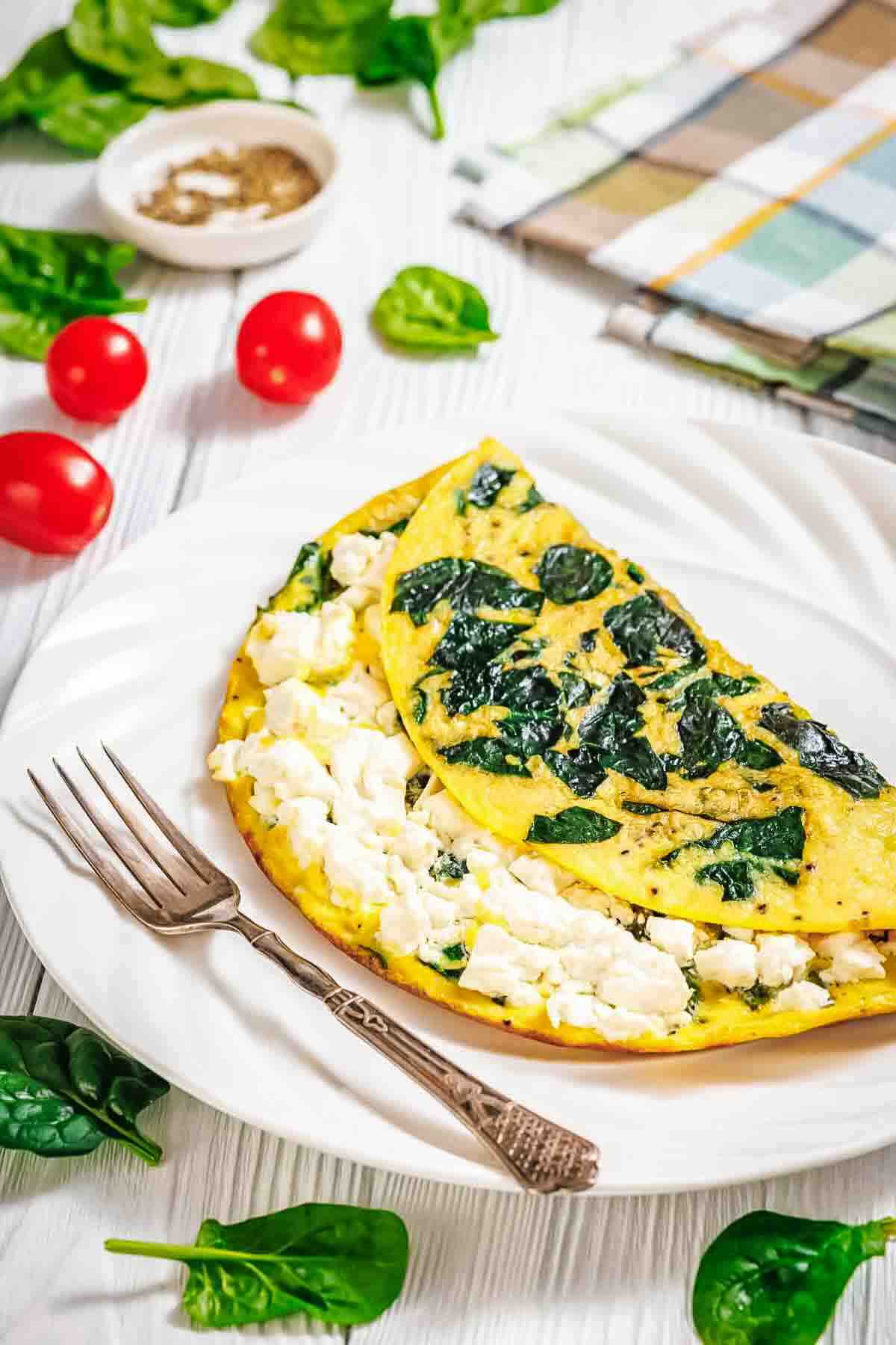 A spinach and feta omelette is served on a white plate with a fork, surrounded by fresh spinach leaves and cherry tomatoes.