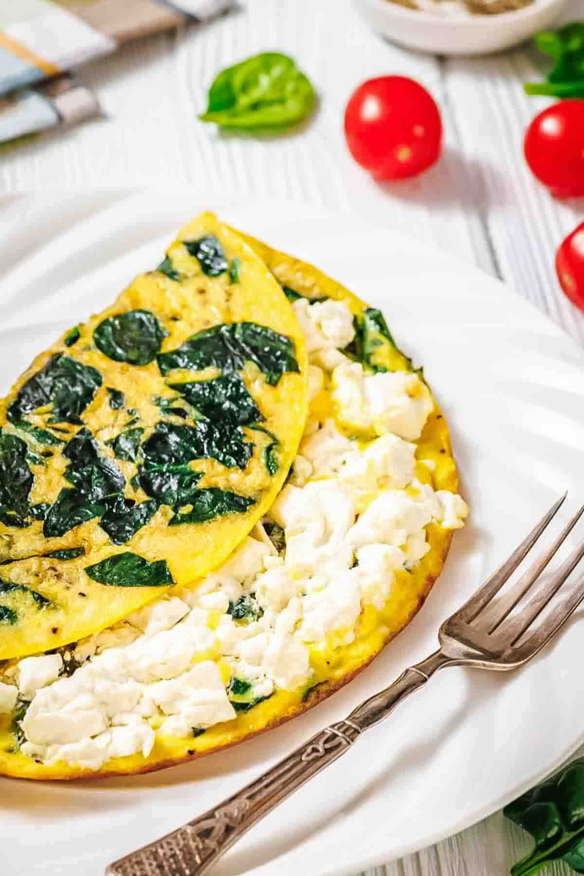 An omelette filled with crumbled feta cheese and spinach is served on a white plate with a fork, surrounded by cherry tomatoes and greens.