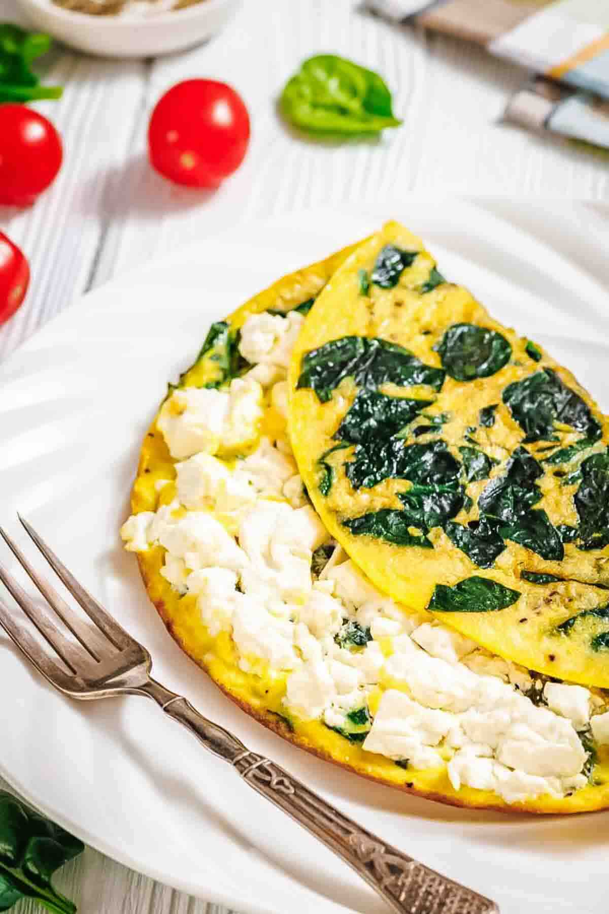 A spinach and feta omelette is served on a white plate with a fork, garnished with cherry tomatoes and spinach leaves in the background.