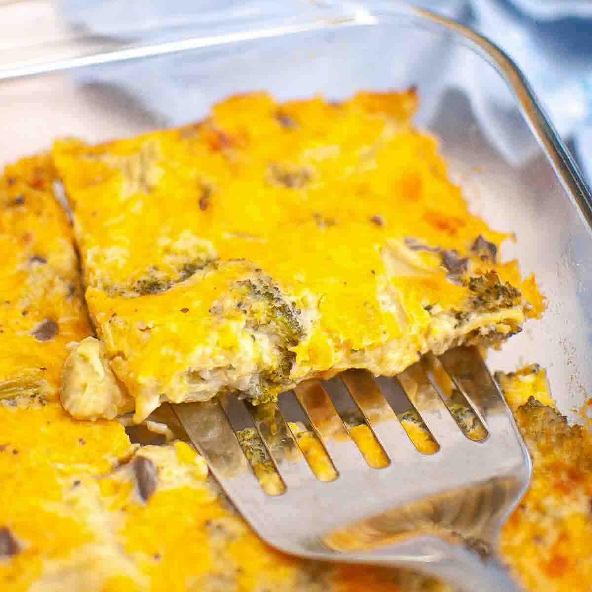 A metal spatula lifts a slice of baked broccoli and cheese casserole from a glass baking dish.