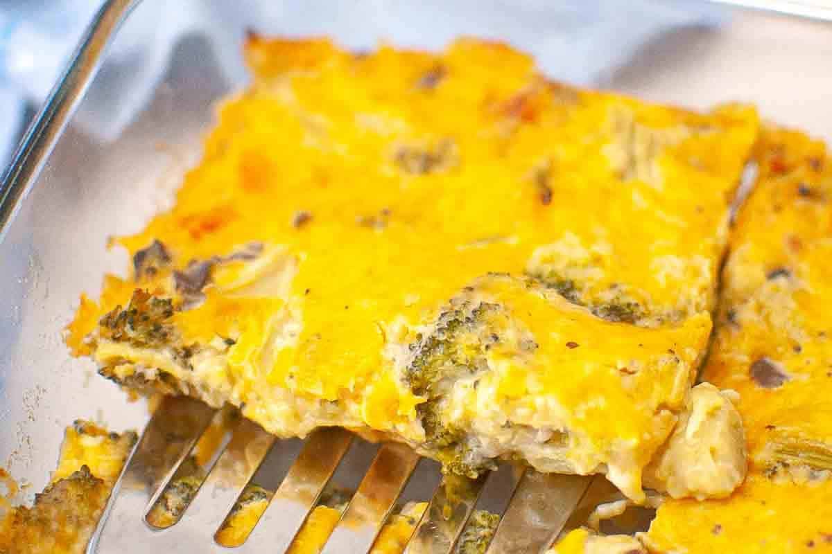 A metal spatula lifts a square piece of baked casserole with cheese, broccoli, and other visible ingredients from a glass baking dish.