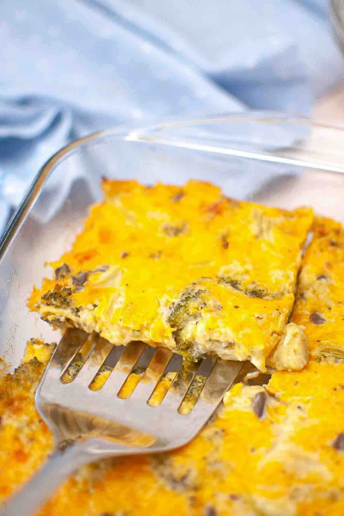 A metal spatula lifts a square piece of baked egg casserole with cheese and vegetables from a glass baking dish.