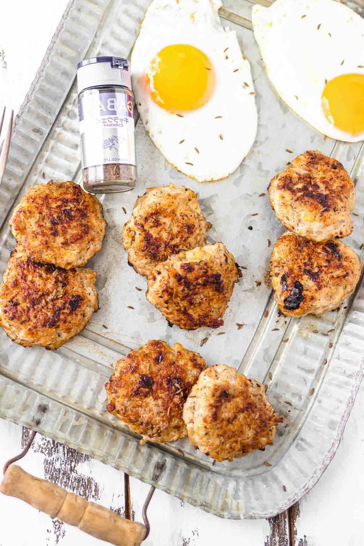 A metal tray with seven browned sausage patties, two sunny-side-up eggs, and a container of seasoning.
