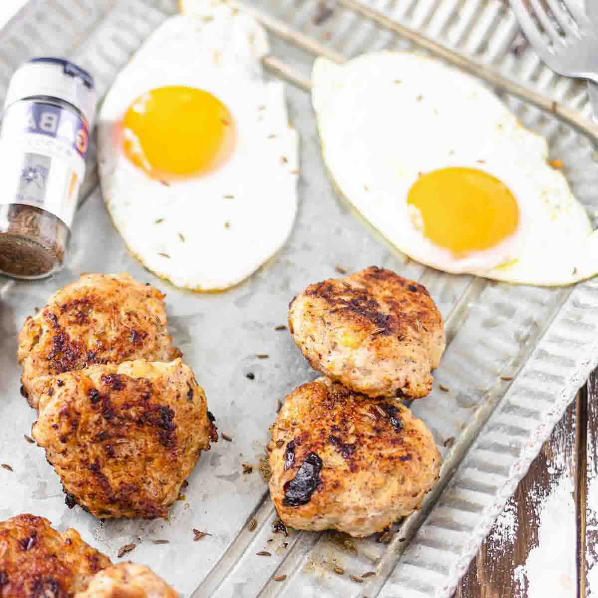 Four browned sausage patties, two sunny-side-up eggs, and a container of black pepper are arranged on a metal tray.