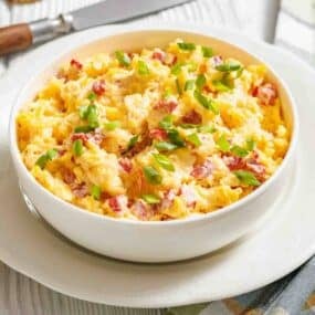 A white bowl filled with scrambled eggs mixed with diced tomatoes and topped with chopped green onions, placed on a white plate.