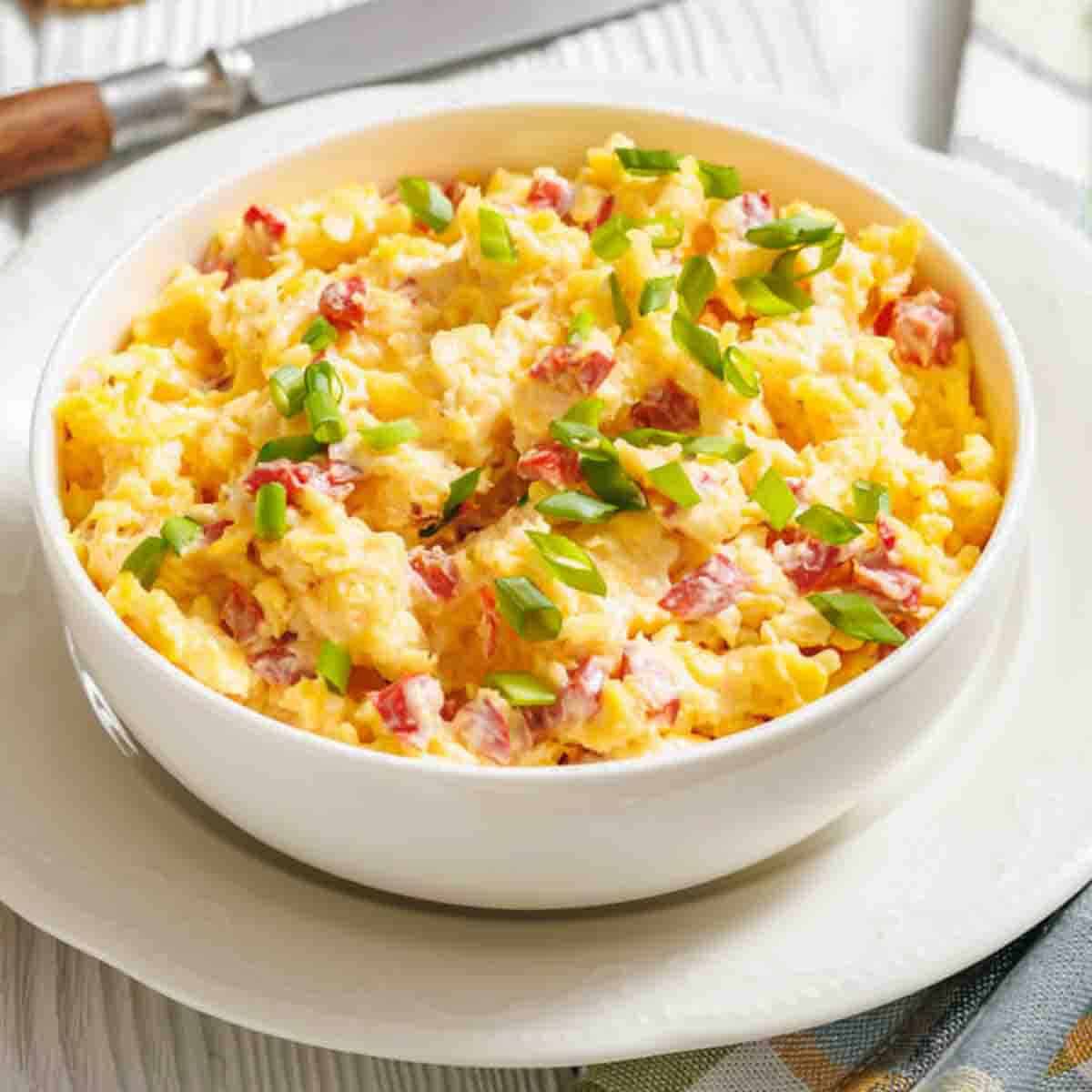 A white bowl filled with scrambled eggs mixed with diced tomatoes and topped with chopped green onions, placed on a white plate.