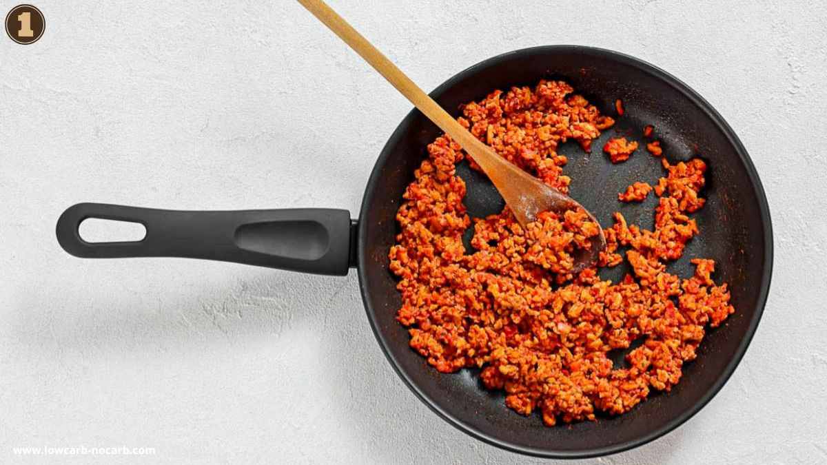 A black frying pan with cooked ground meat in a red sauce, stirred with a wooden spoon, sits on a white surface.