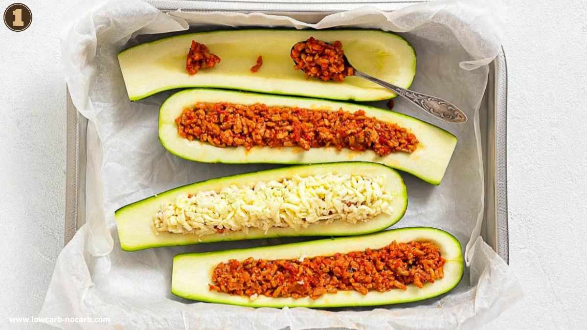 Four zucchini halves on a baking tray lined with parchment paper, hollowed out and filled with ground meat sauce; one is topped with shredded cheese.