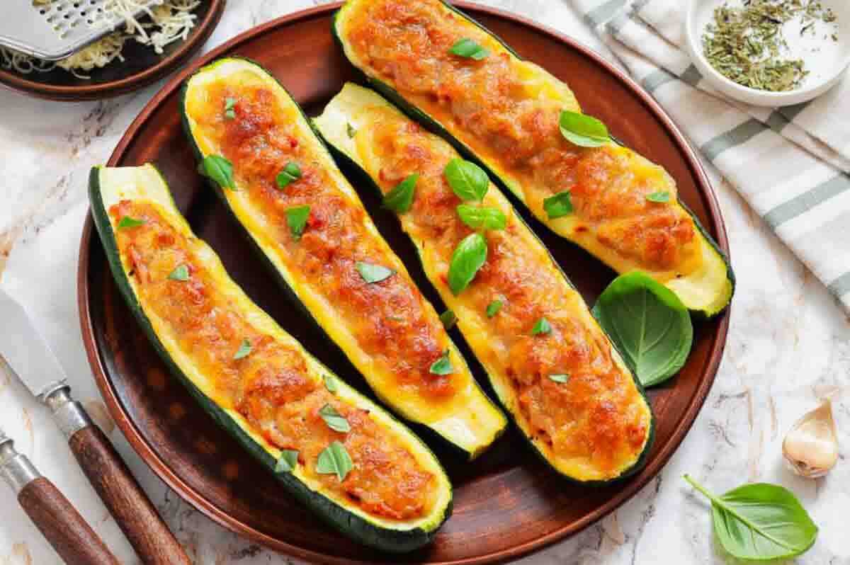 Four zucchini boats filled with a cheesy, baked stuffing are arranged on a brown plate, garnished with fresh basil leaves.