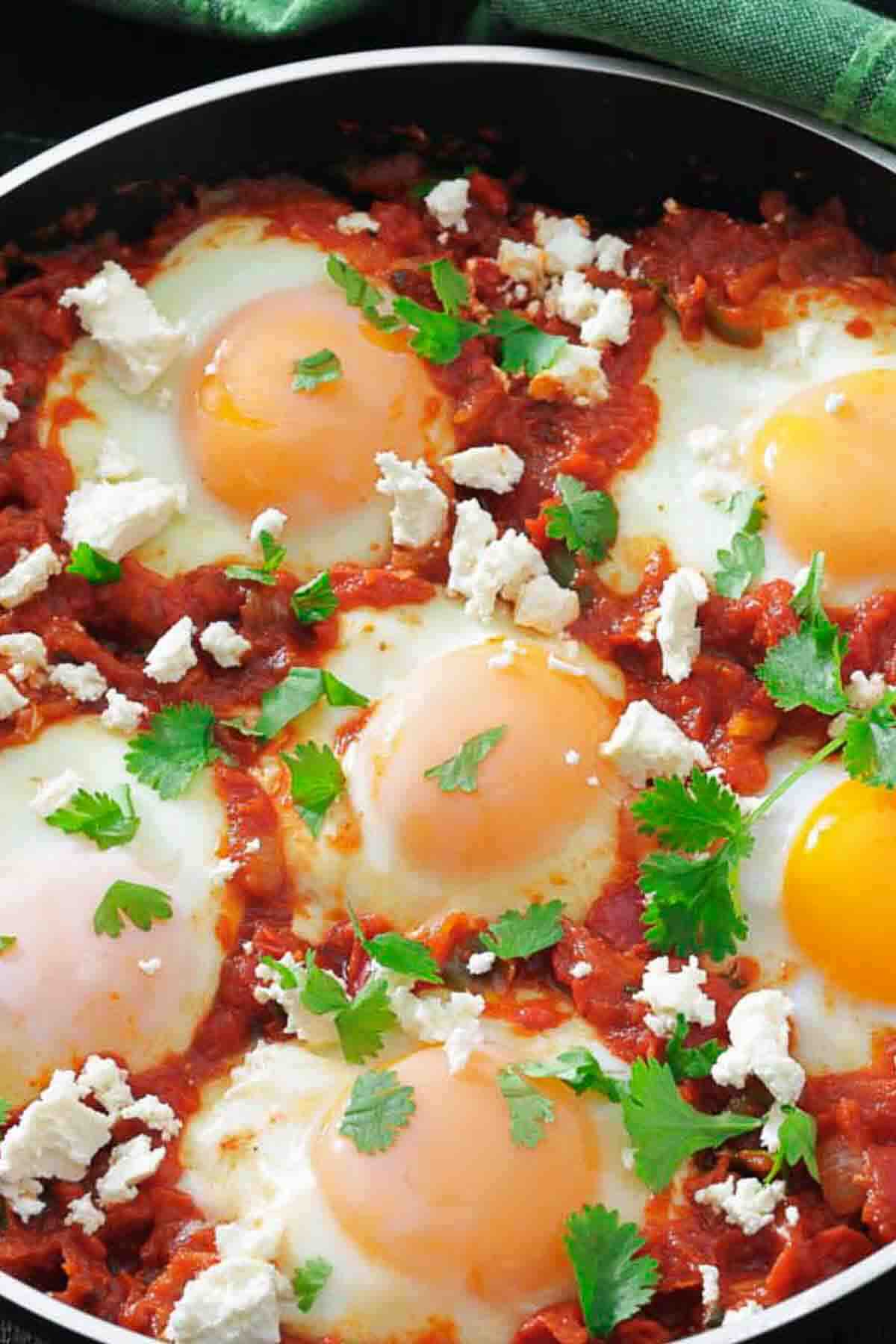 Five eggs cooked in tomato sauce, garnished with crumbled cheese and fresh cilantro, in a skillet.