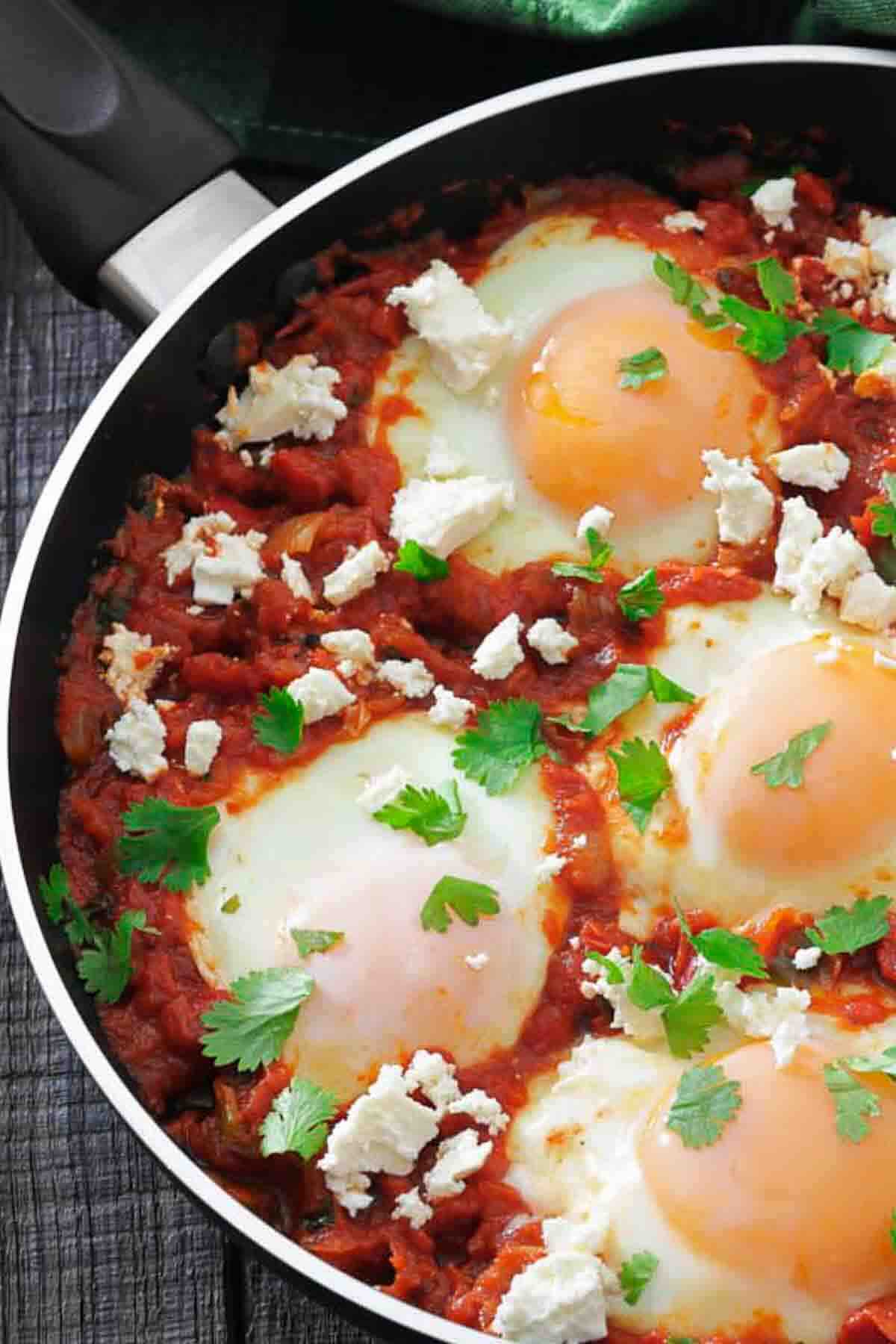 A skillet containing three baked eggs in a tomato sauce, garnished with crumbled cheese and fresh parsley.