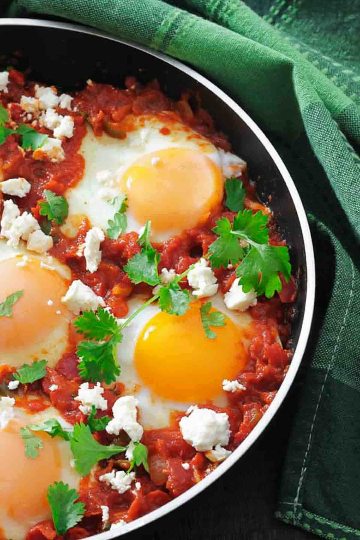 A skillet with three sunny-side-up eggs cooked in a tomato sauce, topped with crumbled cheese and fresh cilantro, next to a green cloth.