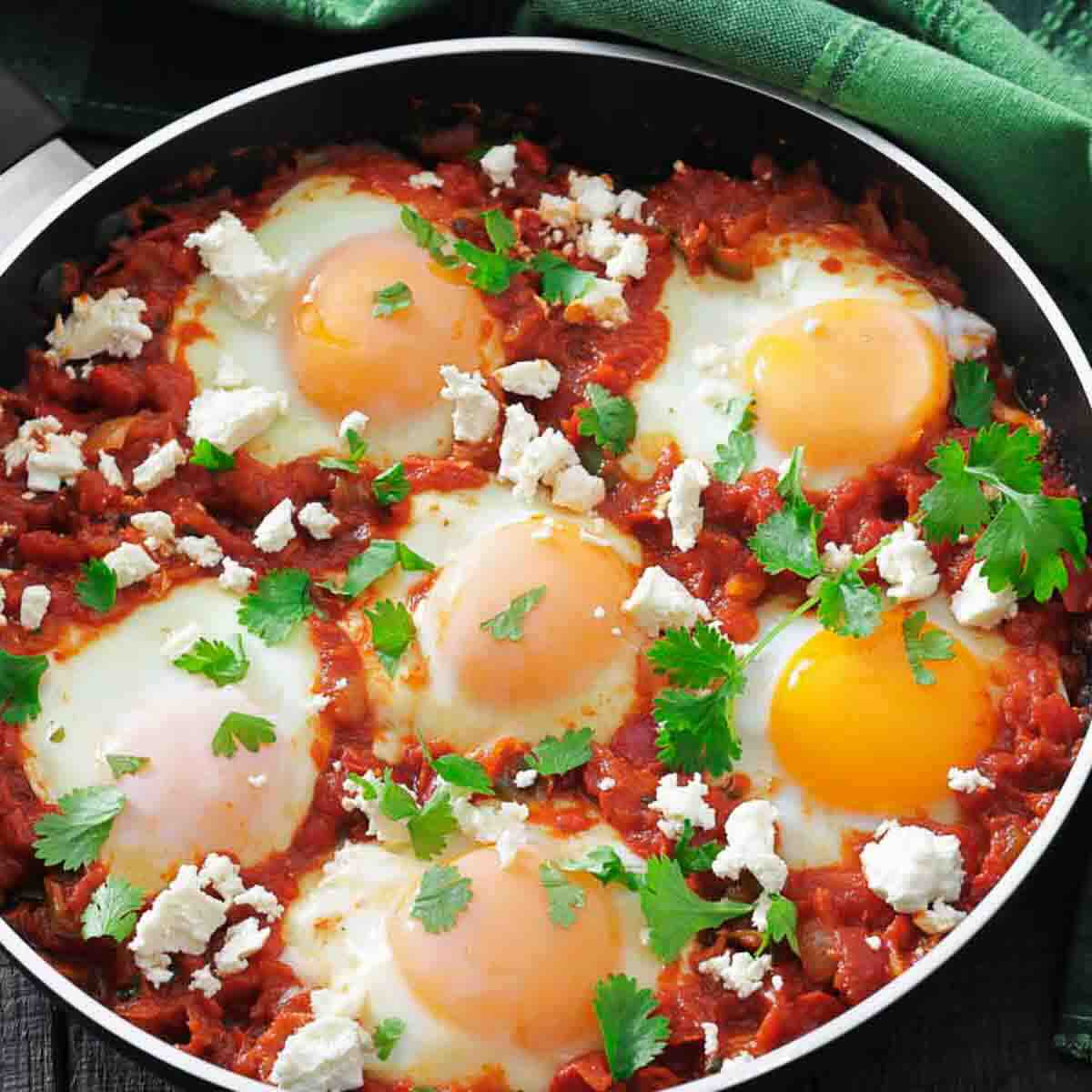 A skillet filled with shakshuka, featuring poached eggs in a tomato sauce, topped with crumbled cheese and fresh parsley.