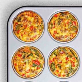 Four baked egg muffins with vegetables and sausage in a metal muffin tin, viewed from above.