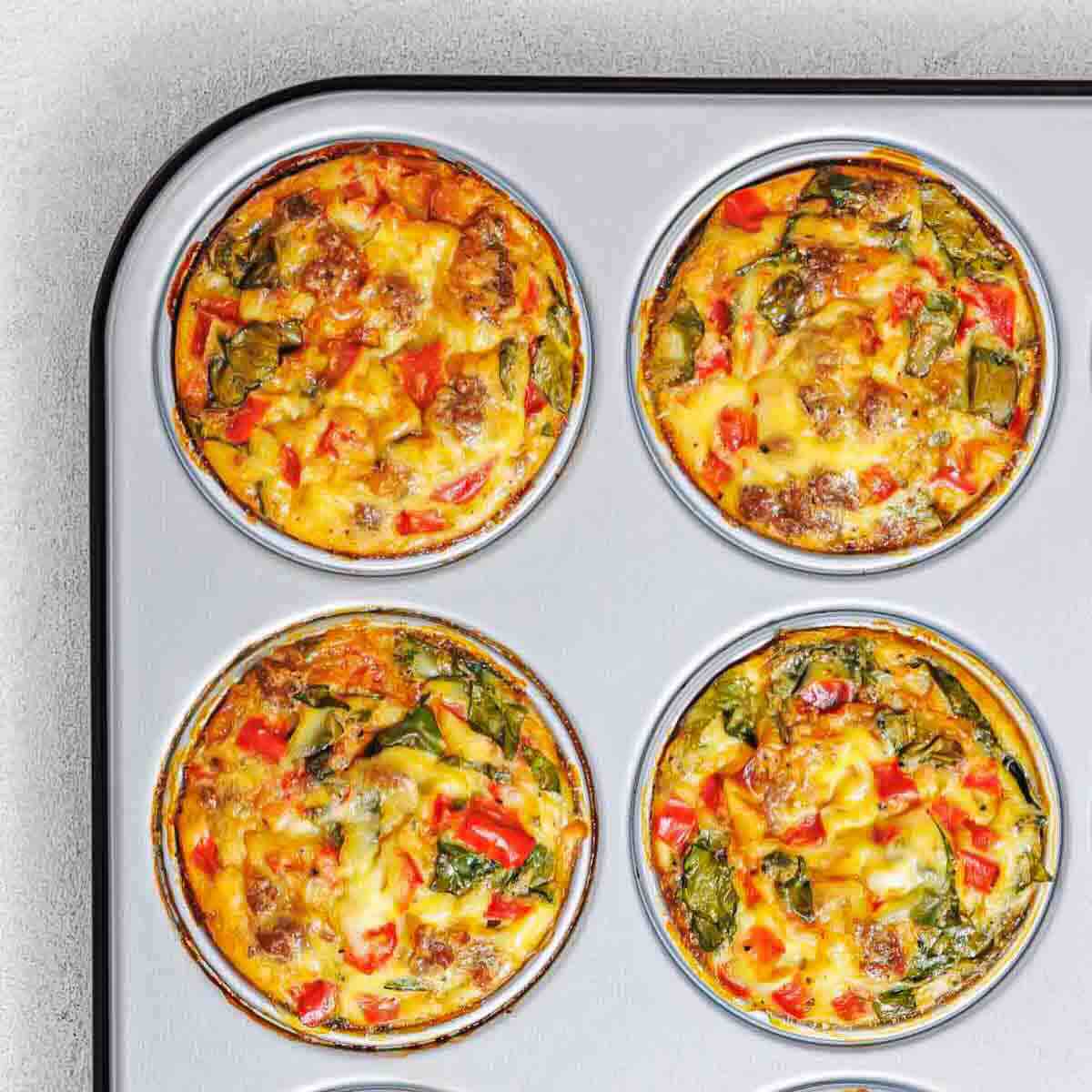 Four baked egg muffins with vegetables and sausage in a metal muffin tin, viewed from above.