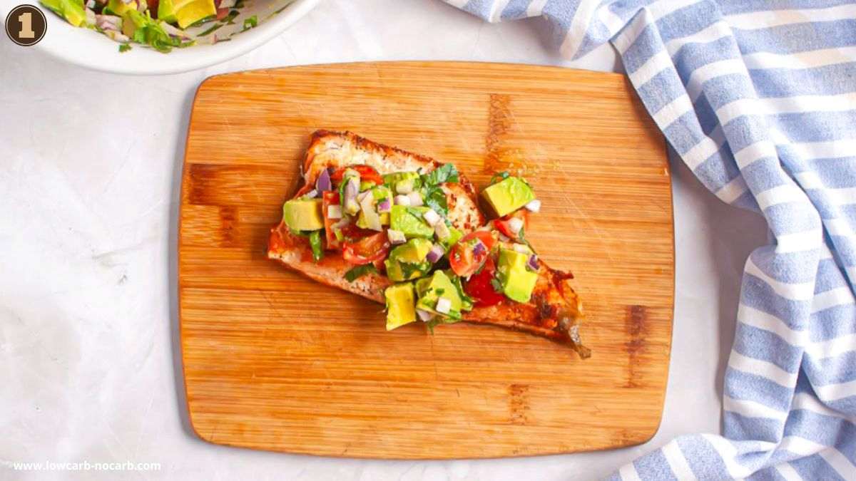 A piece of cooked fish topped with diced avocado, tomatoes, and herbs sits on a wooden cutting board next to a blue and white striped cloth.
