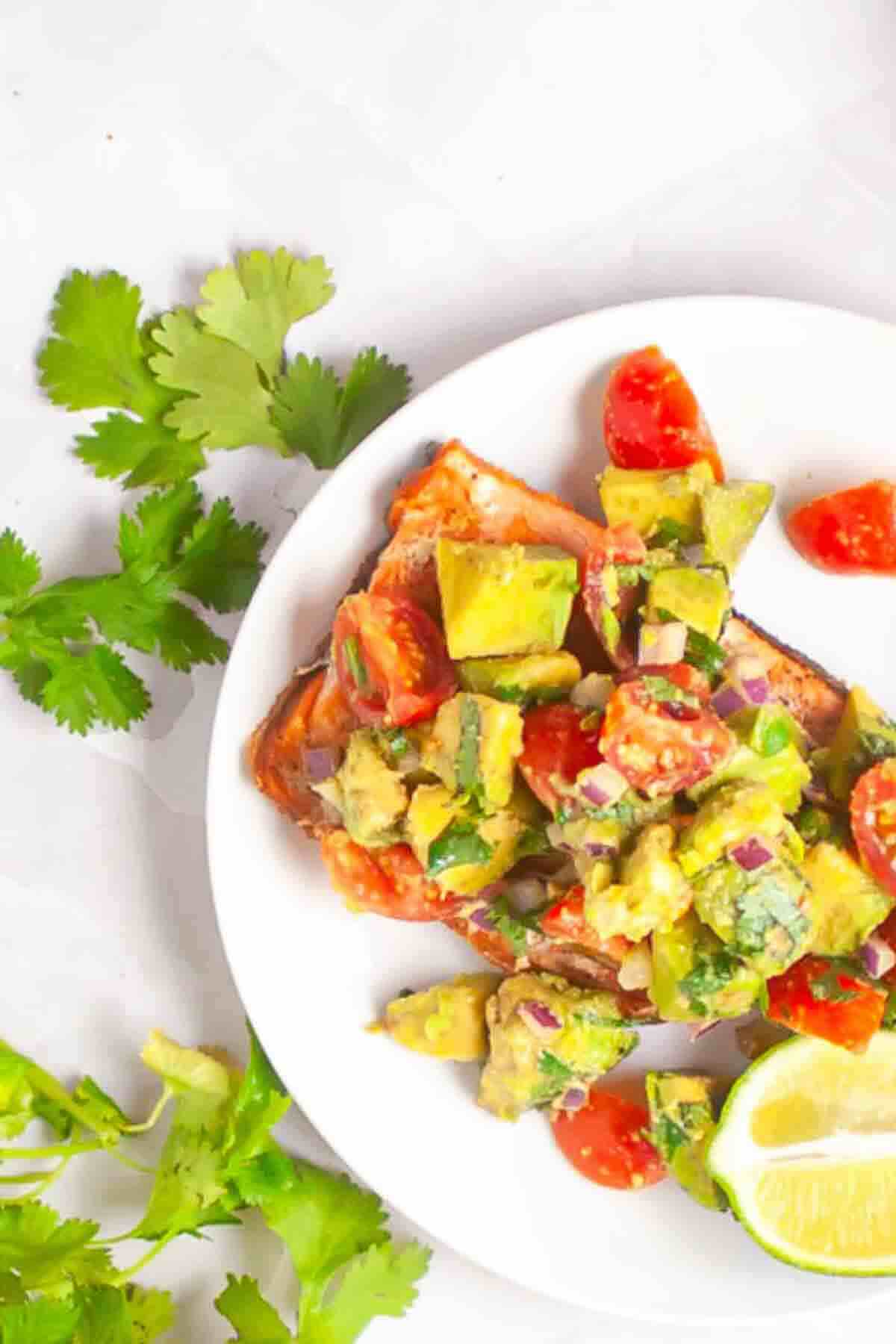 A plate with salmon topped with avocado and tomato salsa, garnished with lime wedges and fresh cilantro on the side.