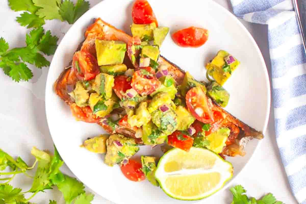 Grilled salmon topped with avocado, tomato, and onion salsa, served with a lime wedge on a white plate.