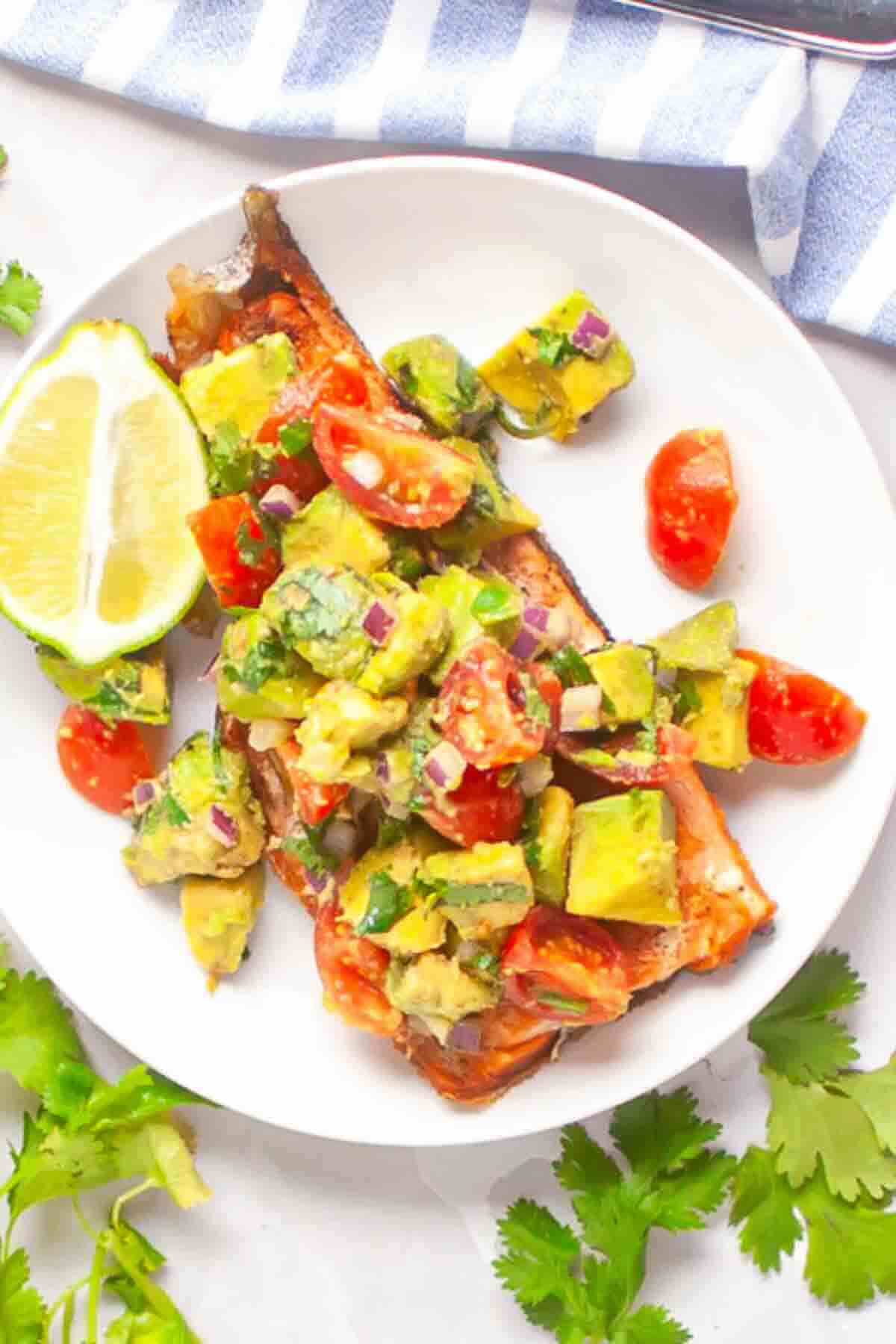 A plate with baked salmon topped with avocado, tomato, and red onion salsa, served with a lime wedge.