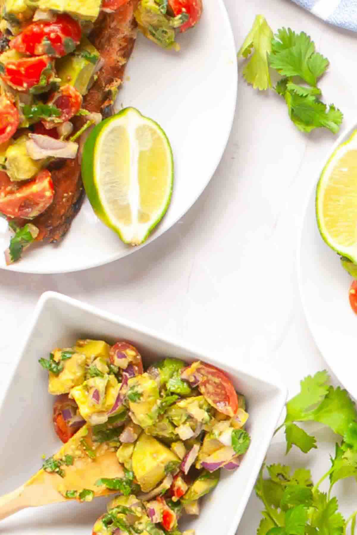 Plates and a bowl with avocado salad containing tomatoes, red onion, and cilantro, served with lime wedges; fresh cilantro leaves on the table.