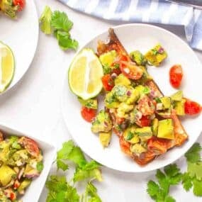 A plate with grilled fish topped with avocado salsa, cherry tomatoes, red onion, and herbs, garnished with lime wedges and fresh cilantro.