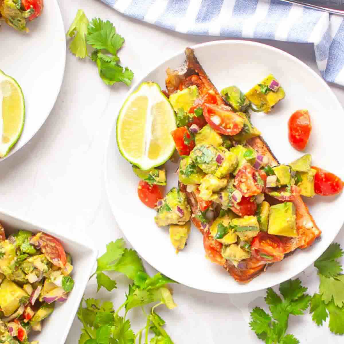 A plate with grilled fish topped with avocado salsa, cherry tomatoes, red onion, and herbs, garnished with lime wedges and fresh cilantro.