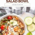 A bowl of buffalo chicken salad with cherry tomatoes, spinach, feta cheese, and sliced cucumbers on the side, set on a white surface with a striped napkin.
