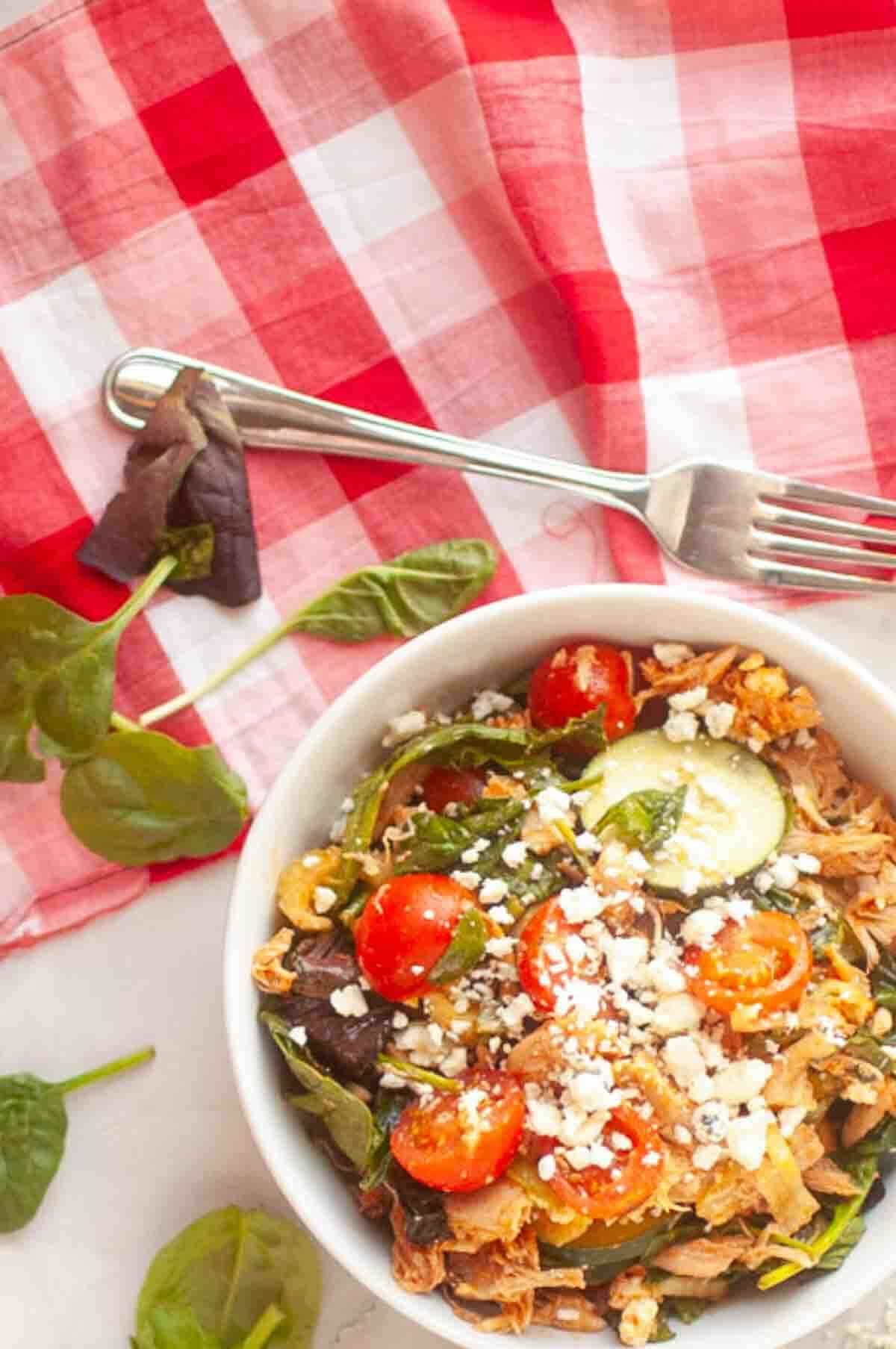 A bowl of salad with cherry tomatoes, cucumbers, spinach, shredded meat, and crumbled feta cheese sits on a white surface next to a red plaid napkin and a fork.