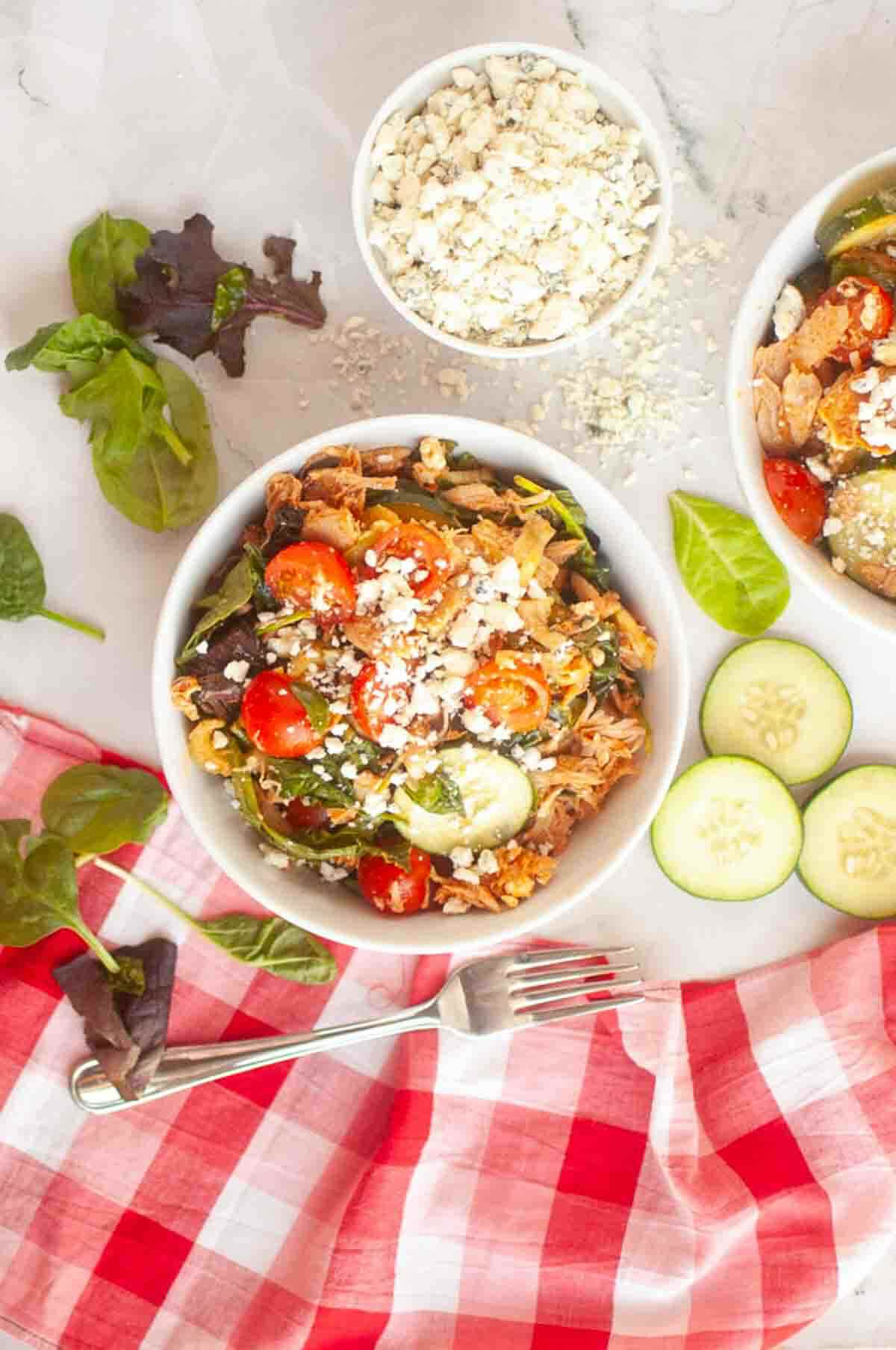 A bowl of salad with cherry tomatoes, cucumbers, greens, shredded chicken, and crumbled cheese, next to sliced cucumbers, loose greens, a fork, and a bowl of cheese on a red checkered cloth.