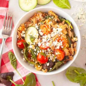 A bowl of salad with shredded chicken, grape tomatoes, cucumber, leafy greens, and crumbled cheese sits on a white surface next to a fork.