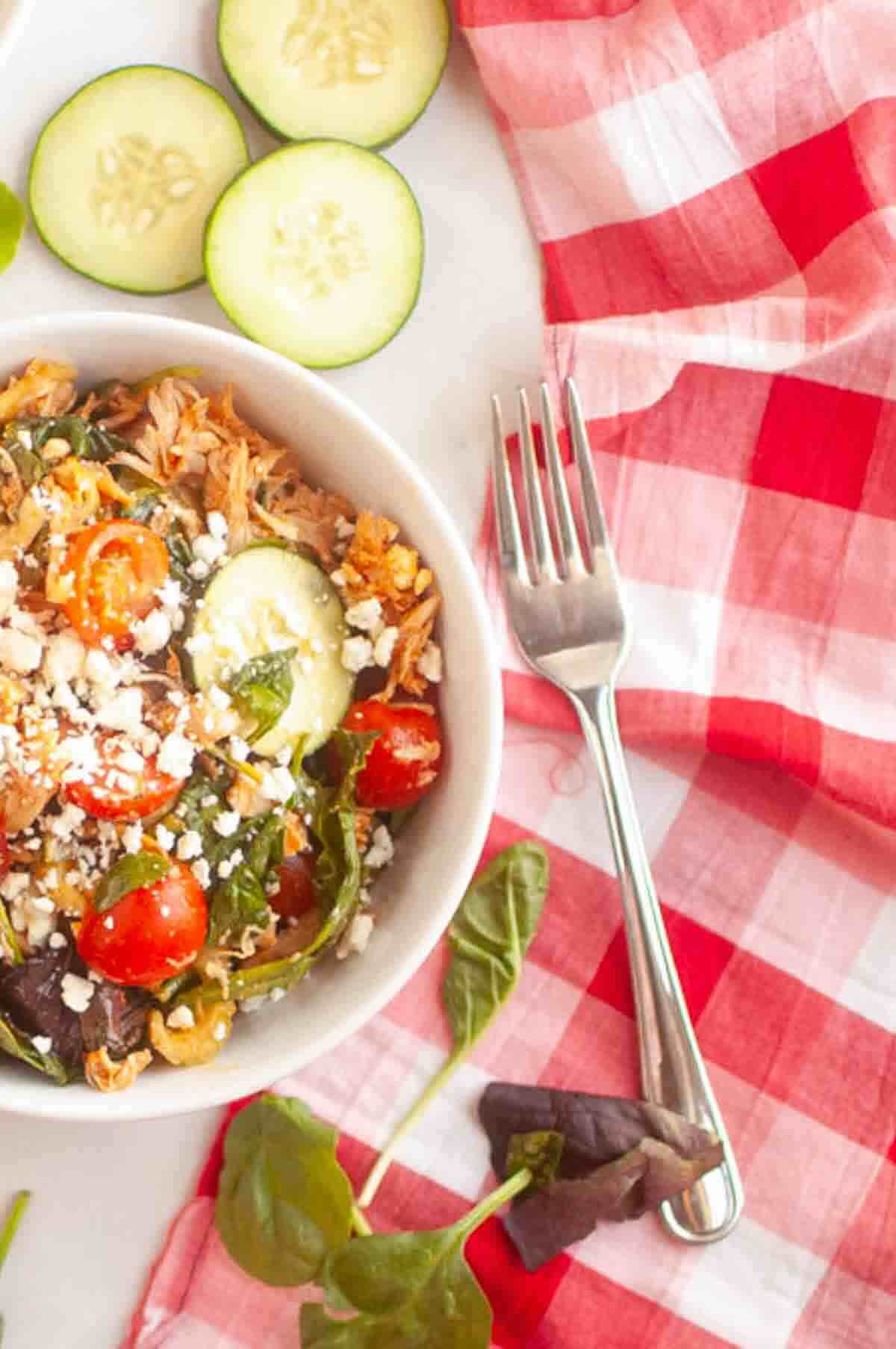 A bowl of salad with cherry tomatoes, cucumbers, greens, and crumbled cheese on a white surface with a fork and a red plaid napkin.
