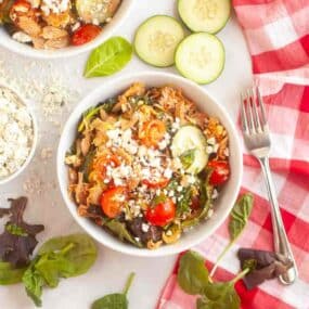 A bowl of salad with shredded chicken, cherry tomatoes, cucumber slices, spinach, and crumbled cheese, placed on a white surface with a red checkered napkin and a fork nearby.