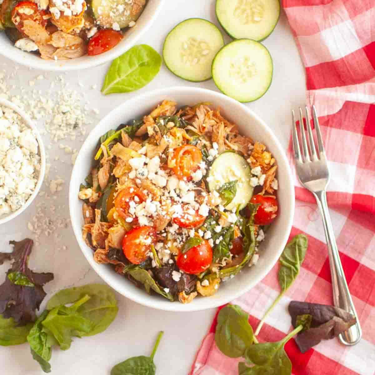 A bowl of salad with shredded chicken, cherry tomatoes, cucumber slices, spinach, and crumbled cheese, placed on a white surface with a red checkered napkin and a fork nearby.