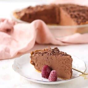 A slice of chocolate pie on a white plate with two raspberries, with the rest of the pie and a pink cloth in the background.
