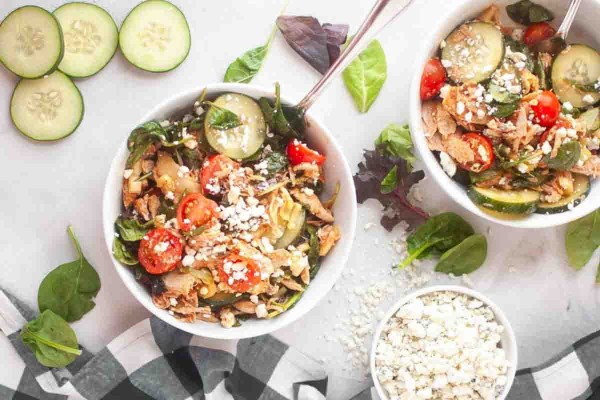 Two bowls of salad with shredded chicken, spinach, cherry tomatoes, cucumber slices, and feta cheese, next to loose greens and a small bowl of extra feta.