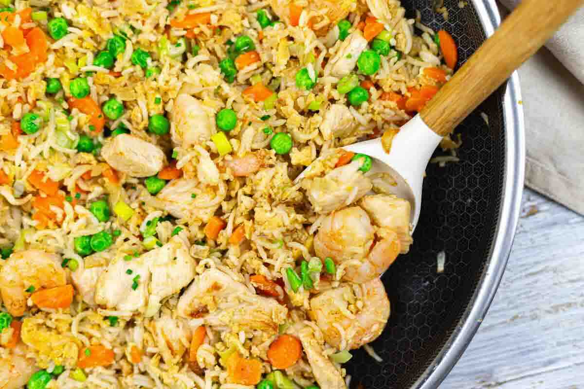 A close-up of fried rice in a pan with shrimp, chicken, carrots, peas, and a serving spoon.