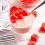 A glass dessert dish filled with pink watermelon sorbet topped with diced watermelon pieces, with a spoon and scattered watermelon cubes nearby.