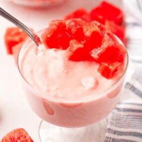 A glass dish filled with pink watermelon mousse, topped with diced watermelon pieces, sits on a white surface next to a striped cloth.