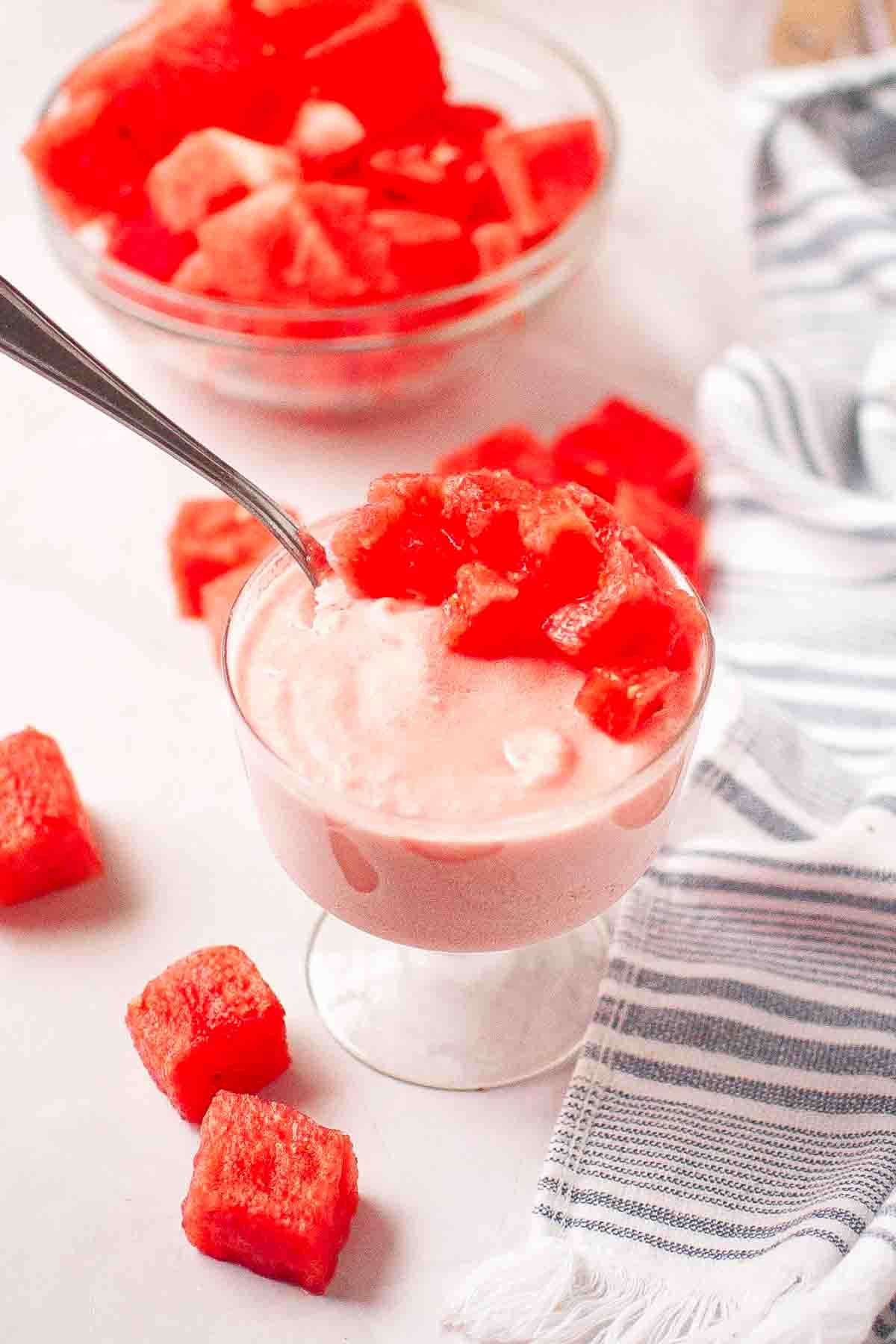 A glass dessert cup filled with pink ice cream or sorbet, topped with watermelon cubes, with more watermelon pieces and a striped towel nearby.